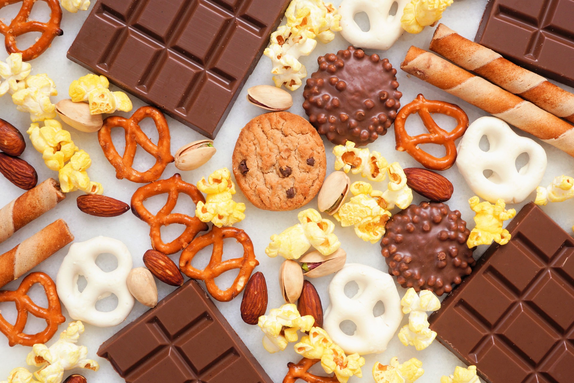 Background of various sweet and salty snacks including chocolate, popcorn, pretzels, nuts and cookies